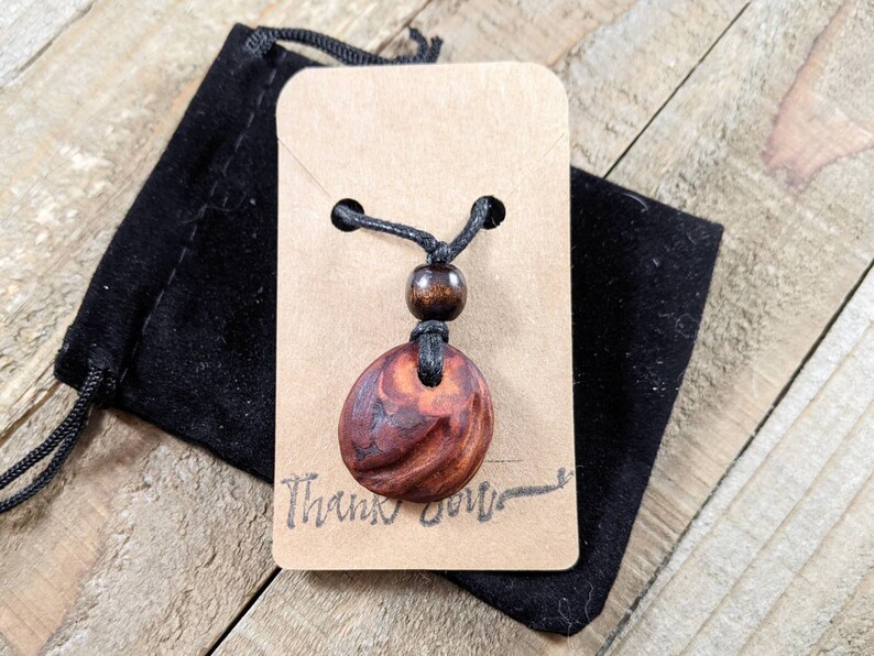 May include: A wooden pendant necklace with a dark brown, oval-shaped pendant. The pendant hangs from a black cord with a wooden bead. The necklace is displayed on a brown tag with the words "Thank you" and a black velvet pouch.