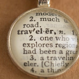 May include: A silver charm necklace with a glass pendant featuring a vintage dictionary definition of the word "traveler". The definition reads: "2. one who visits and explores regions that had been a great distance from home."