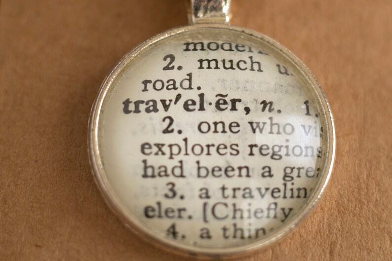 May include: A silver charm necklace with a glass pendant featuring a vintage dictionary definition of the word "traveler". The definition reads: "2. one who visits and explores regions that had been a great distance from home."