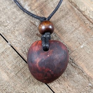 May include: A dark red, heart-shaped pendant necklace. The pendant is made of wood and has a natural grain pattern. It hangs from a black cord with a wooden bead accent.