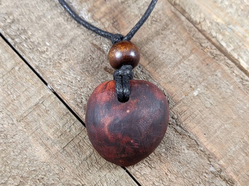 May include: A dark red, heart-shaped pendant necklace. The pendant is made of wood and has a natural grain pattern. It hangs from a black cord with a wooden bead accent.