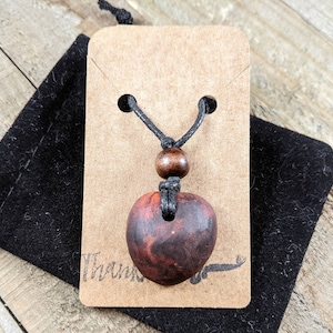 May include: A heart-shaped pendant necklace with a dark wood grain finish. The pendant hangs from a black cord with a wooden bead accent. The necklace is displayed on a brown tag and a black velvet pouch.