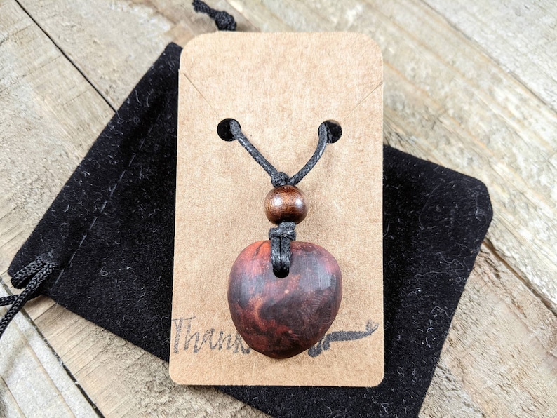 May include: A heart-shaped pendant necklace with a dark wood grain finish. The pendant hangs from a black cord with a wooden bead accent. The necklace is displayed on a brown tag and a black velvet pouch.