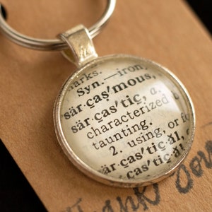 May include: A silver keychain with a glass charm featuring a dictionary definition of the word "sarcastic". The definition reads: "Syn. iron sarcastic, sarcastic, a. characterized by taunting. 2. using, or for sarcastic al, cas'tical"