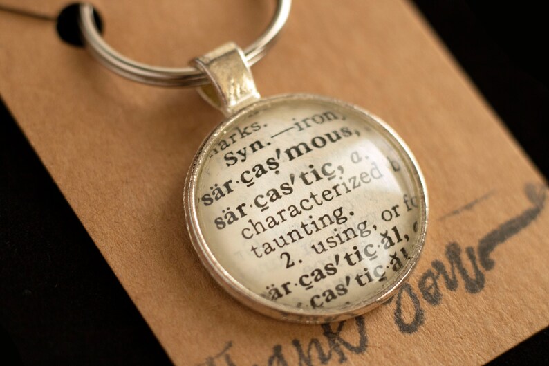 May include: A silver keychain with a glass charm featuring a dictionary definition of the word "sarcastic". The definition reads: "Syn. iron sarcastic, sarcastic, a. characterized by taunting. 2. using, or for sarcastic al, cas'tical"