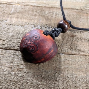 May include: A handcrafted wooden pendant necklace. The pendant is a reddish-brown color with a carved spiral design. It hangs from a black cord with a wooden bead and a sliding knot for adjustment. The necklace is displayed on a wooden surface.