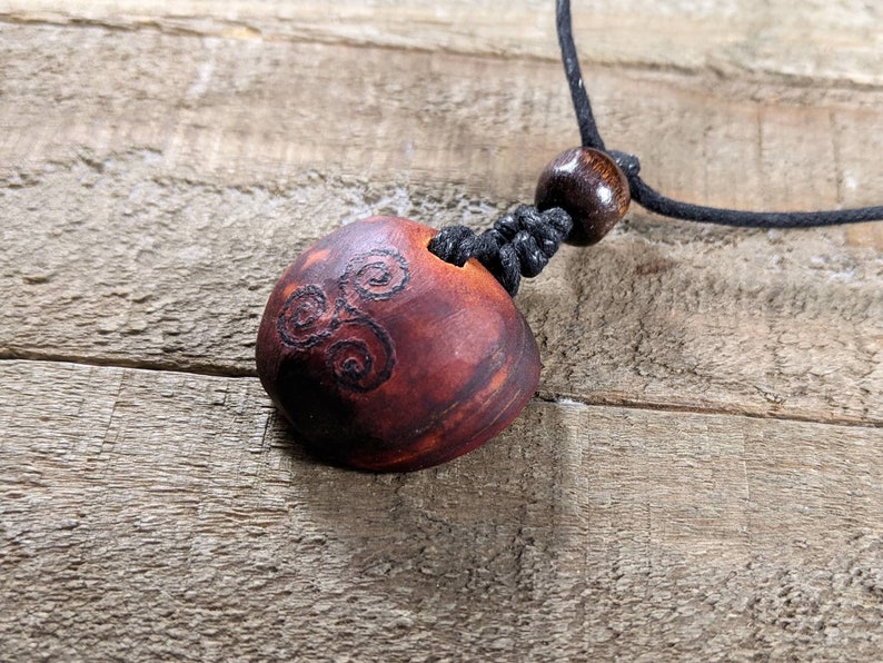 May include: A handcrafted wooden pendant necklace. The pendant is a reddish-brown color with a carved spiral design. It hangs from a black cord with a wooden bead and a sliding knot for adjustment. The necklace is displayed on a wooden surface.