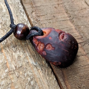 May include: A dark brown wooden pendant with a natural, irregular shape and a black cord. The pendant has a reddish-brown hue and a textured surface.