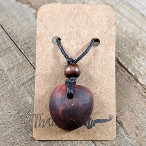 May include: A handcrafted wooden pendant necklace with a heart-shaped pendant. The pendant is a deep reddish-brown color, suspended from a black cord with a wooden bead accent. The necklace is displayed on a brown card.