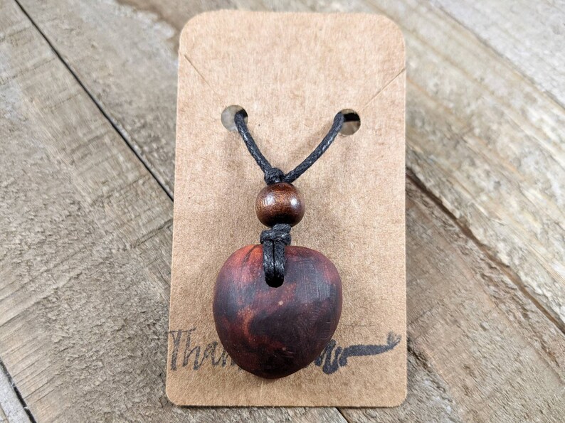 May include: A handcrafted wooden pendant necklace with a heart-shaped pendant. The pendant is a deep reddish-brown color, suspended from a black cord with a wooden bead accent. The necklace is displayed on a brown card.