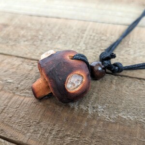 May include: A brown ceramic mushroom pendant with clear quartz accents hangs from a black cord with a brown wooden bead.