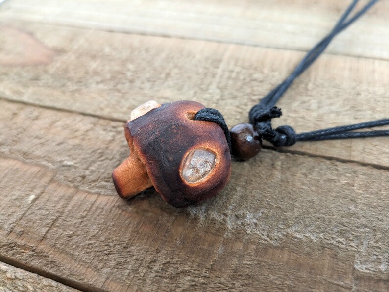 May include: A brown ceramic mushroom pendant with clear quartz accents hangs from a black cord with a brown wooden bead.
