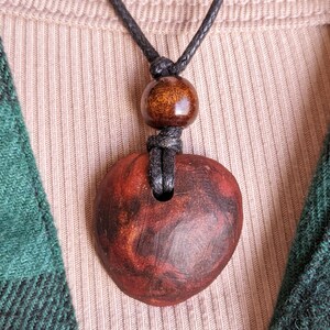 May include: A handmade wooden pendant necklace. The pendant is a reddish-brown color with dark brown marbling. The necklace has a black cord with a wooden bead above the pendant. The background includes a beige ribbed shirt and a green and black plaid shirt.