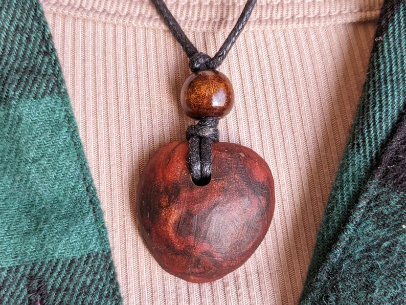 May include: A handmade wooden pendant necklace. The pendant is a reddish-brown color with dark brown marbling. The necklace has a black cord with a wooden bead above the pendant. The background includes a beige ribbed shirt and a green and black plaid shirt.
