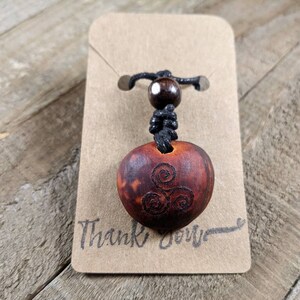 May include: A wooden pendant necklace with a carved Celtic spiral design. The pendant is a reddish-brown color and is attached to a black cord with a wooden bead. The pendant is displayed on a brown card with the words "Thank You" written on it.