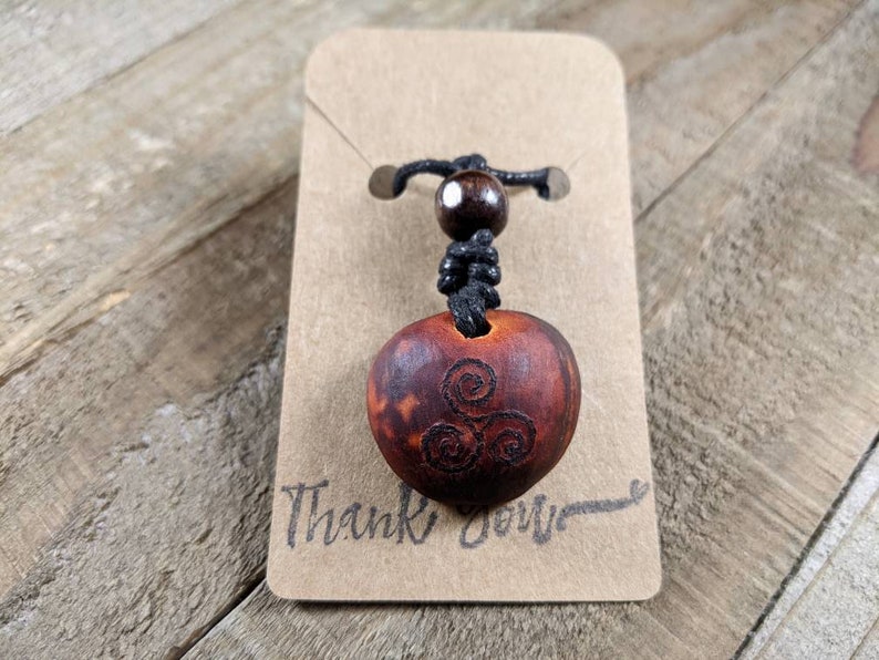 May include: A wooden pendant necklace with a carved Celtic spiral design. The pendant is a reddish-brown color and is attached to a black cord with a wooden bead. The pendant is displayed on a brown card with the words "Thank You" written on it.