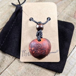 May include: A wooden pendant necklace with a carved spiral design. The pendant is a reddish-brown color and is suspended from a black cord. The necklace is displayed on a brown card with the word "Thank" visible. A black pouch is behind the card.