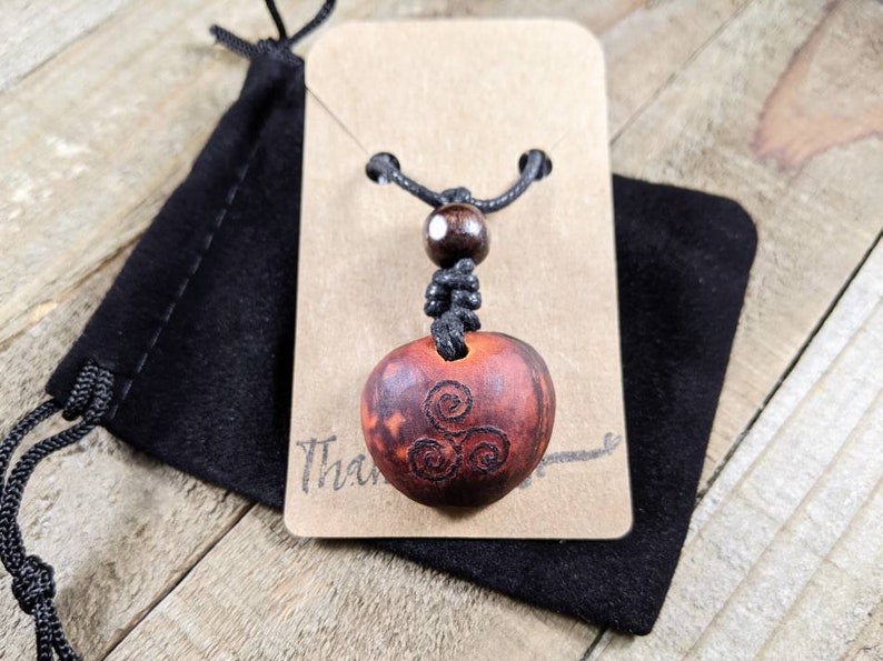 May include: A wooden pendant necklace with a carved spiral design. The pendant is a reddish-brown color and is suspended from a black cord. The necklace is displayed on a brown card with the word "Thank" visible. A black pouch is behind the card.