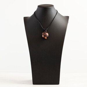 May include: A brown wooden mushroom pendant necklace on a black cord. The pendant is on a black mannequin bust.