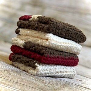 May include: A stack of four crocheted dishcloths in brown, cream and burgundy. The dishcloths are folded and stacked on a wooden surface.