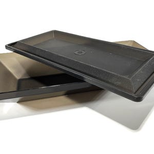 May include: A black plastic rectangular tray with a matching lid. The tray has a slightly raised edge and the lid has a small circular indentation.