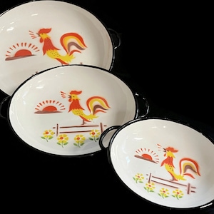 May include: Three white enamelware bowls with black rims and rooster designs. Each bowl features a rooster with a red comb and yellow feathers, a sunrise, and a fence with flowers.