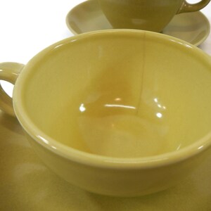 May include: A yellow ceramic teacup and saucer set. The cup has a smooth, glossy finish and a wide, rounded shape. The saucer is a matching yellow and has a slightly raised rim.