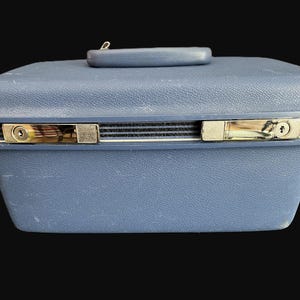 May include: A vintage blue suitcase with a handle on top and two metal latches. The suitcase has a textured surface and a black trim around the middle. The latches are gold-colored.