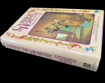 May include: A book titled "Camber the Heretic" by Katherine Kurtz. The book cover features an illustration of a scene from the book, with two figures in a room. The book is part of the "Legends of Camber of Culda" series.
