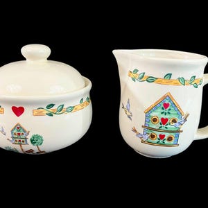 May include: A white ceramic sugar bowl and creamer set with a birdhouse design. The set features a green birdhouse with red hearts and blue birds, surrounded by green vines and yellow accents.