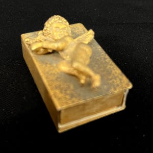 May include: A rectangular gold-toned metal box featuring a three-dimensional relief of a winged cherub figure resting on the lid. The surface of the box has a mottled, weathered texture with a metallic finish, and the lid is hinged.