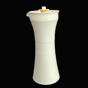 May include: A tall, white ceramic pitcher with a flared base and a narrow neck. It has a spout and a small, yellow-toned lid. The pitcher's design is modern and elegant, suitable for serving beverages.