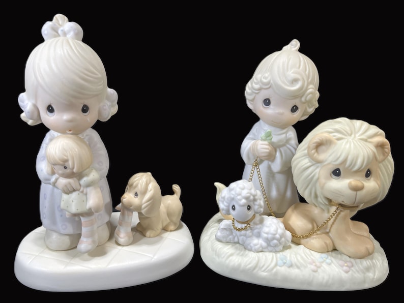 Puede incluir: Dos figuritas de porcelana de personajes de Precious Moments. La primera figurita es una ni&ntilde;a que sostiene una mu&ntilde;eca. La segunda figurita es un ni&ntilde;o que sostiene un cordero. Ambas figuritas est&aacute;n sobre bases blancas.
