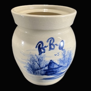 May include: Off-white ceramic BBQ jar with a blue landscape design. The jar features the word "B-B-Q" in blue script above a rural scene with trees, a building, and birds. The jar has a wide opening.