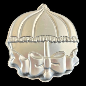 May include: A silver metal cake pan shaped like an umbrella with a bow. The pan is designed for baking a cake in the shape of an umbrella.