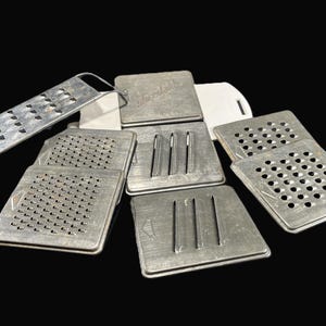 May include: A set of eight silver metal kitchen graters with different sized holes and patterns. The graters are arranged in a pile on a white surface.