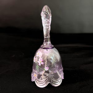 May include: A clear glass bell with a purple tint, featuring a detailed handle and a floral design. The bell has a scalloped bottom edge and a painted floral pattern with pink and purple flowers.