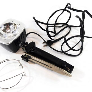 May include: A black and silver photographic flash unit with a wire cage, a black cord, and a black clamp.