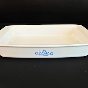 May include: A rectangular white ceramic baking dish with a smooth, glossy finish. The exterior front center features a small, detailed blue floral pattern consisting of three stylized cornflowers with leaves and stems centered on the side wall.