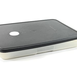 May include: A clear plastic rectangular storage container with a black lid. The lid has a small white circle in the centre.