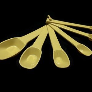 May include: A set of six yellow plastic measuring spoons. The spoons are arranged in a fan shape, with the largest spoon at the top and the smallest spoon at the bottom.