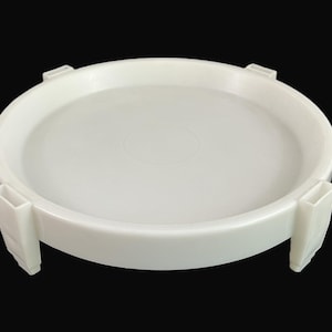 May include: A white, round plastic tray with a raised edge and four rectangular supports. The tray is designed with a shallow interior and is set against a black background.