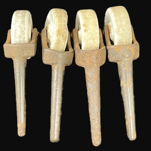 May include: A set of four vintage-style furniture casters, each featuring a weathered, dark brown cast iron stem and a worn, off-white wheel. The metal stems show a textured, oxidized surface, while the wheels appear to be made of a hard, aged material, all arranged in a row against a dark background.