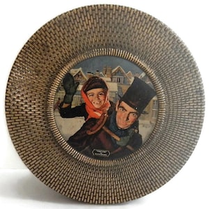 May include: A round, gold-colored plate with a raised, textured rim. The center of the plate features a framed image of a man and a child in a snowy winter scene. The man is wearing a top hat and the child is wearing a red scarf and mittens.