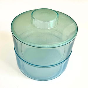 May include: A two-piece, light blue glass jar with a lid. The jar has a cylindrical shape with a slightly flared base. The lid is a lighter shade of blue and has a small, rounded handle. The jar is likely for storage.