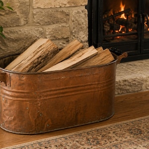 May include: An oval-shaped copper log holder with two side handles featuring black grips. The container has a metallic finish and is filled with several pieces of split firewood. The structure is made of hammered copper with visible rivets.