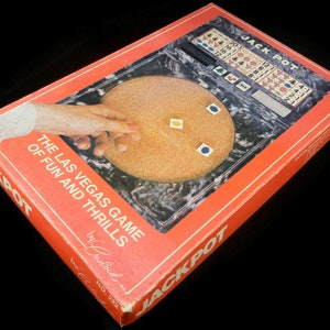 May include: A red and white box for the "Jackpot" Las Vegas game of fun and thrills by Crisloid. The box features a marbleized surface with a spinning wheel and dice.