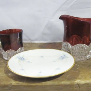 May include: A set of three vintage glass pieces, including a red glass creamer, a red glass cup, and a white saucer with a blue floral design.