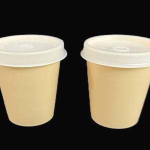 May include: Two disposable paper cups with a light beige color and a smooth finish. Each cup is topped with a circular, white plastic lid that features a small, centered drinking opening. The cups have a tapered shape and a flat base.