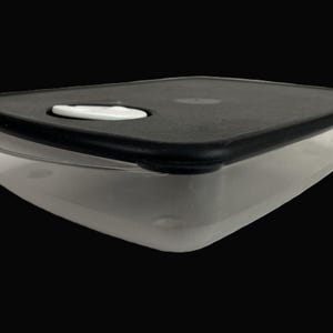 May include: A clear, rectangular food storage container with a black lid. The lid has a white, oval-shaped button in the center. The container is made of clear plastic and is designed for food storage.